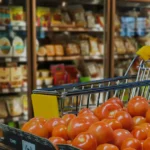 Shopping cart filled with fresh tomatoes in a grocery store aisle with shelves of products in the background.