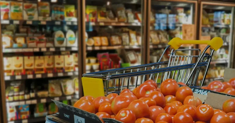 Shopping cart filled with fresh tomatoes in a grocery store aisle with shelves of products in the background.