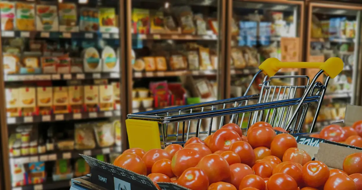 Shopping cart filled with fresh tomatoes in a grocery store aisle with shelves of products in the background.