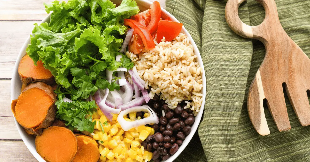 Lettuce, sweet potato, rice, tomato, corn, black beans, onion in a white bowl next to a wooden fork.